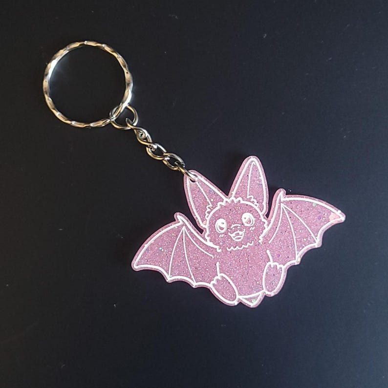 May include: A pink glittery bat keychain featuring a cartoon-style bat with large ears and outstretched wings. The bat has white line art details and is attached to a silver-colored metal split ring by a short chain link, creating a decorative accessory.