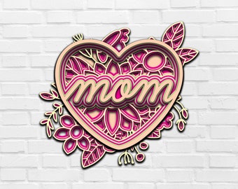 May include: A pink and white layered paper cut design of a heart with the word "mom" inside. The heart is surrounded by floral designs.