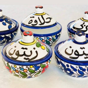 Może przedstawiać: Zestaw pięciu białych ceramicznych miseczek z niebieskimi i kolorowymi wzorami kwiatowymi. Każda miseczka ma pokrywkę z arabską kaligrafią. Miseczki są oznaczone jako "Halawa", "Zbib", "Shouf", "Rish" i "Misk".