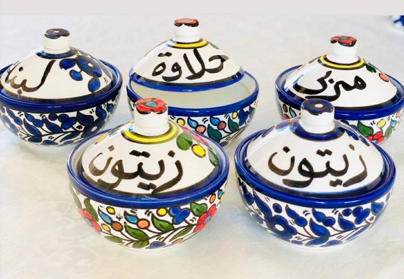 Może przedstawiać: Zestaw pięciu białych ceramicznych miseczek z niebieskimi i kolorowymi wzorami kwiatowymi. Każda miseczka ma pokrywkę z arabską kaligrafią. Miseczki są oznaczone jako "Halawa", "Zbib", "Shouf", "Rish" i "Misk".