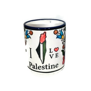 May include: White ceramic mug with a blue rim and a colorful floral design. The mug features a stylized outline of Palestine with the Palestinian flag colors and the text "I Love Palestine".
