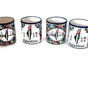 May include: Five ceramic mugs with a white background and blue trim. Each mug features a colorful design of the Palestinian flag and the words "I Love Palestine". The mugs are arranged in a row on a white background. The image also includes a logo for "Ceramic Store Art" with a stylized olive branch and floral design.