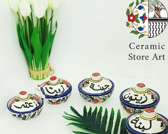 May include: Five small ceramic bowls with lids, each decorated with colorful floral patterns and Arabic calligraphy. The bowls are arranged on a white surface with a green leafy plant in the background.