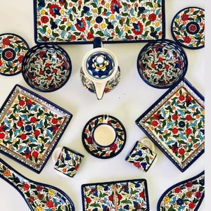 Może przedstawiać: Zestaw kolorowych ceramicznych naczyń z kwiatowymi wzorami w odcieniach niebieskiego, czerwonego, ż&oacute;łtego i zielonego. Zestaw zawiera talerze, miski, kubki, czajnik i łyżkę.
