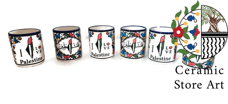 May include: Five ceramic mugs with blue and white trim. Each mug features a colorful design of the Palestinian flag and the words "I Love Palestine" in English and Arabic. The mugs are arranged in a row on a white background.