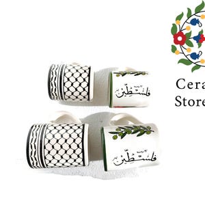 May include: Four ceramic mugs with different designs. Two mugs feature a black and white geometric pattern, one mug has a green olive branch design, and one mug has a green olive branch design with Arabic text that reads "Felesteen".