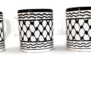 May include: Three white ceramic mugs with black geometric patterns. The mugs have black rims and handles. The mugs are arranged in a row from left to right. The image also includes a logo for "Ceramic Store Art" with a stylized tree and floral design.