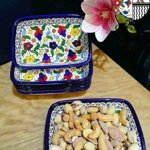 Puede incluir: Un juego de cuatro platos rectangulares de cerámica azul y blanca con un patrón floral. Los platos están apilados uno encima del otro. El plato superior está lleno de una variedad de nueces, incluyendo anacardos, almendras y pistachos.