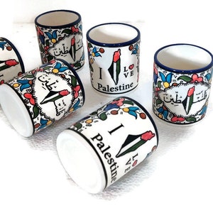 May include: Set of five white ceramic mugs with blue trim. Each mug features a colorful design with the words "I Love Palestine" in English and Arabic. The mugs are arranged in a circle on a white background.
