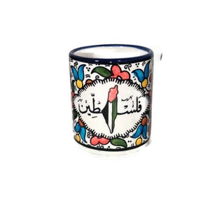 May include: A white ceramic mug with a blue rim and a colorful floral design. The mug features a green, white, and black Palestinian flag with Arabic text that reads "Palestine".