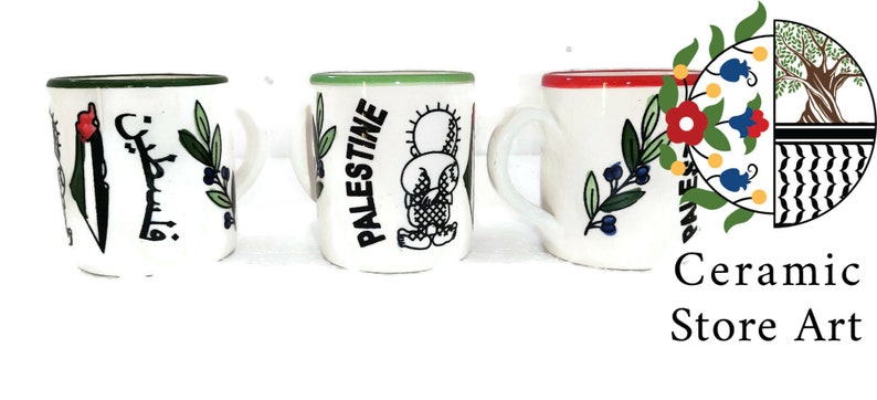 May include: Three white ceramic mugs with different designs. The first mug has a green rim and features a green olive branch, a red flower, and the outline of Palestine in green. The second mug has a green rim and features a green olive branch, a cartoon figure, and the word "Palestine" in black. The third mug has a red rim and features a green olive branch with blue berries and a red flower. The mugs are on a white surface.