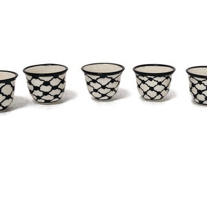 Può includere: Sei piccole tazze in ceramica bianca con motivi geometrici neri e bordi neri. Le tazze sono disposte in fila su uno sfondo bianco. L'immagine include anche un logo con un design in bianco e nero e il testo "Ceramic Store Art".