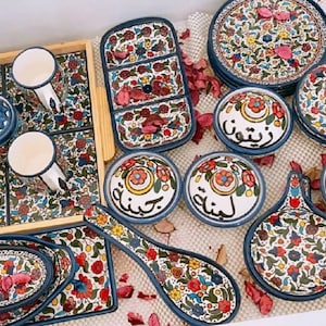 Może przedstawiać: Zestaw kolorowych ceramicznych naczyń z kwiatowymi wzorami i arabską kaligrafią. Naczynia obejmują miski, talerze i łyżkę do serwowania. Naczynia są ułożone na drewnianej tacy.