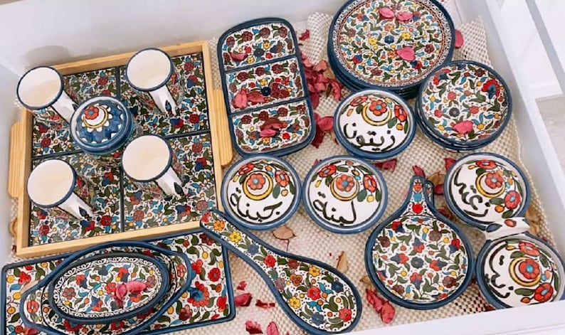 Może przedstawiać: Zestaw kolorowych ceramicznych naczyń z kwiatowymi wzorami i arabską kaligrafią. Naczynia obejmują miski, talerze i łyżkę do serwowania. Naczynia są ułożone na drewnianej tacy.