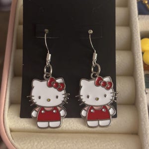 Puede incluir: Un par de pendientes colgantes de Hello Kitty. Cada pendiente presenta un gato blanco con un lazo rojo y un vestido rojo. Los pendientes son de metal plateado y se exhiben en una tarjeta negra. Un accesorio lindo.