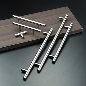 May include: A set of five brushed nickel cabinet pulls in various sizes and styles. The pulls are arranged on a dark brown wooden surface.