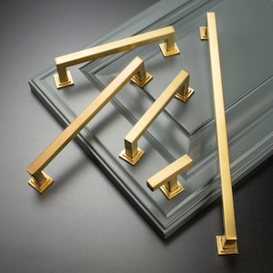 May include: Five gold cabinet pulls with a square design. The pulls are arranged on a gray surface.