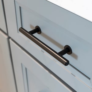 May include: Black cabinet drawer pull with a rectangular bar and round end caps. The pull is mounted on a light blue cabinet door.