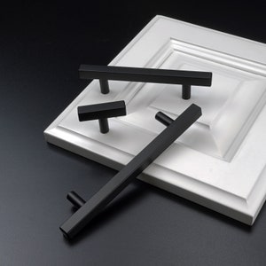 May include: Three black cabinet pulls, two are rectangular and one is a T-shaped pull. The pulls are shown on a white cabinet door.