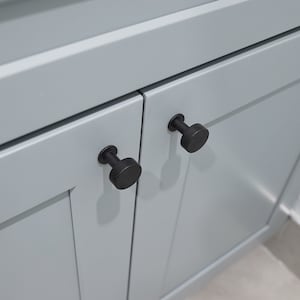 May include: Two black cabinet knobs with a round shape are mounted on a light gray cabinet door.