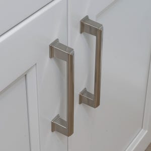 May include: Two brushed nickel cabinet pulls with a rectangular shape and a slightly curved design. The pulls are mounted on a white cabinet door.
