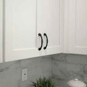 May include: Two black cabinet pulls with a decorative design are mounted on white cabinet doors. The cabinets are above a white tiled backsplash.