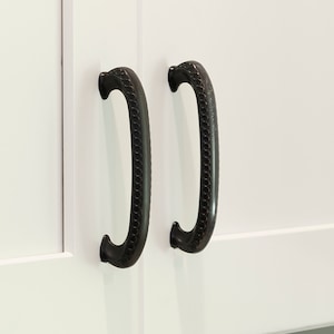 May include: Two black cabinet pulls with a decorative rope design. The pulls are mounted on a white cabinet door.