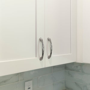 May include: Two white cabinet doors with silver, decorative handles. The cabinets are mounted above a tiled backsplash.