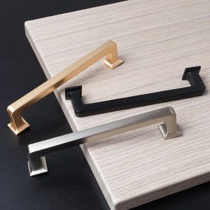 May include: Three cabinet pulls in different finishes: gold, black, and silver. Each pull has a rectangular shape with squared edges.