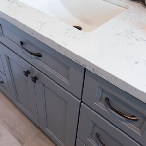 May include: Gray bathroom vanity with white countertop and a white porcelain sink. The vanity has multiple drawers with black hardware.