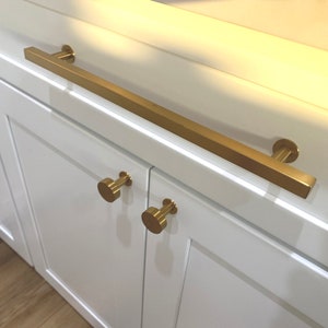 May include: Gold cabinet hardware set with a long bar handle and two round knobs. The hardware is installed on white cabinet doors.