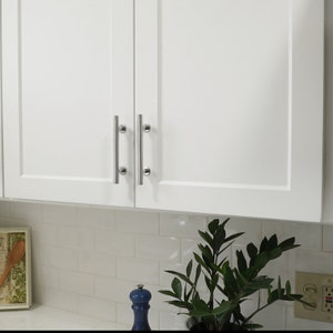May include: White kitchen cabinets with silver bar handles. The cabinets are closed and are above a white tiled backsplash.