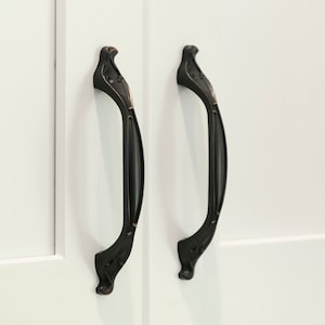 May include: Two black cabinet door pulls with an ornate design. The pulls are made of metal and have a slightly aged finish.
