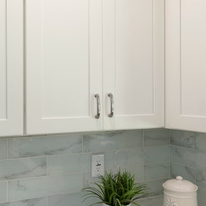 May include: White kitchen cabinets with silver handles above a light gray marble tile backsplash. A small potted plant and a white ceramic jar are on the counter.