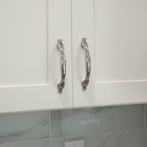May include: Two silver cabinet pulls with an ornate design. The pulls are mounted on white cabinet doors.