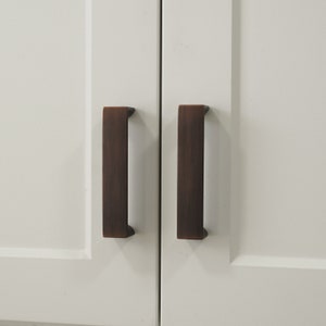 May include: Two cabinet doors with brushed bronze cabinet pulls. The pulls are rectangular and have a slightly rounded edge.