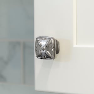 May include: A silver cabinet knob with a square base and an ornate design featuring a central circle and four leaf-like shapes.