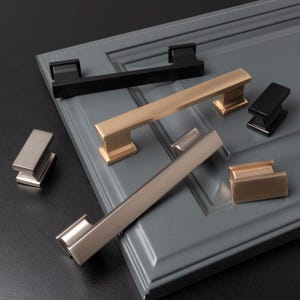 May include: Five different cabinet pulls in various finishes, including black, gold, and silver. The pulls are displayed on a gray surface.