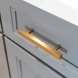 May include: A brushed gold cabinet pull with square ends. The pull is mounted on a light blue cabinet door.