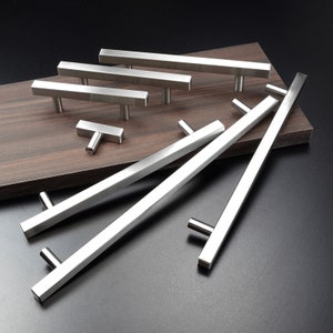 May include: A set of ten silver cabinet pulls in various sizes. The pulls are made of metal and have a brushed finish. The pulls are arranged on a brown wooden surface.