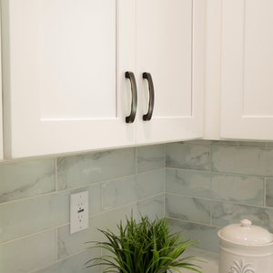 May include: White kitchen cabinets with black arched handles. The cabinets are above a tiled backsplash with a white electrical outlet.