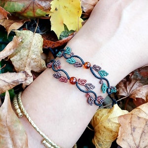 Op de afbeelding: Twee zwarte koordarmbanden met groene en oranje kralen, die op bladeren lijken, op een pols tegen een achtergrond van gevallen herfstbladeren.