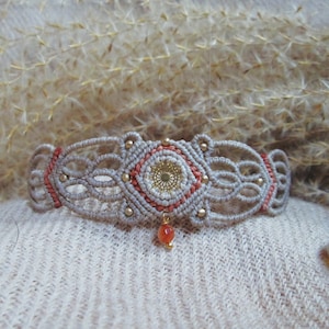 Puede incluir: Un brazalete de macramé beige con una pieza central roja y dorada y un colgante de piedra roja.