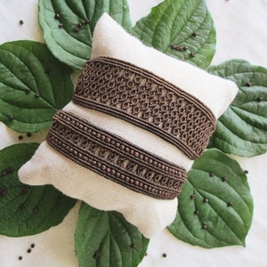 Puede incluir: Dos pulseras de cuentas marrones con un diseño tejido sobre un fondo de tela blanca. Las pulseras están dispuestas sobre una almohada blanca con hojas verdes alrededor.