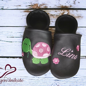 Könnte beinhalten: Ein Paar schwarze Lederschlappen mit Stickereien. Ein Schlappen zeigt eine grüne Schildkröte mit rosa Punkten und der andere hat den Namen "Lina" in rosa gestickt.