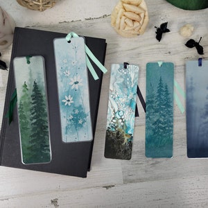 May include: Five different bookmarks with various designs. The bookmarks are all rectangular and have a ribbon attached to the top. The designs include a watercolor painting of trees in a foggy forest, a watercolor painting of white flowers, a blue and white abstract design, a watercolor painting of trees in a forest, and a watercolor painting of a foggy forest.