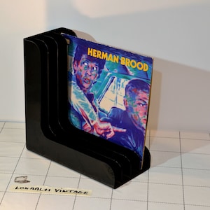 Op de afbeelding: Zwarte plastic platenrek met drie vakken. Een vinylplaat met de titel "Herman Brood" wordt in het rek getoond.