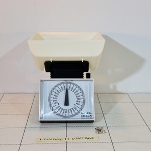 May include: A white kitchen scale with a black dial and a white bowl. The dial has markings for grams and kilograms. The scale is on a tiled surface.