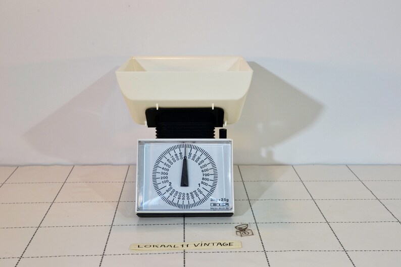 May include: A white kitchen scale with a black dial and a white bowl. The dial has markings for grams and kilograms. The scale is on a tiled surface.