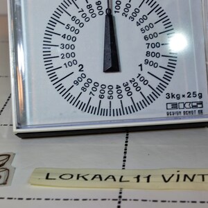May include: White kitchen scale with a black dial and needle. The dial has markings for grams and kilograms. The scale reads 3kg x 25g. The text "EKS DESIGN OCHOT SK" is printed on the scale.
